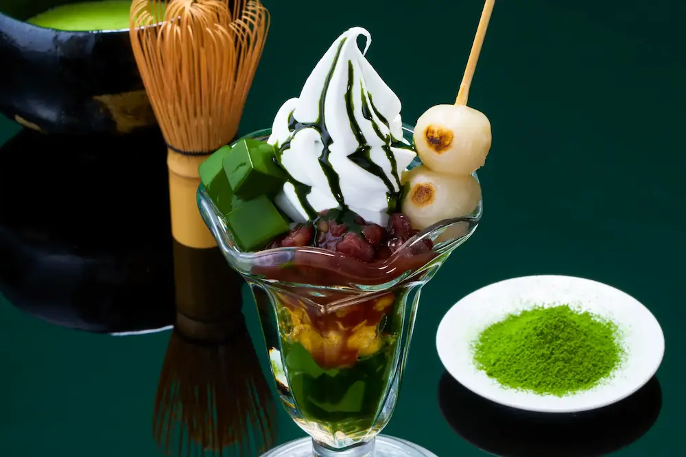 串団子、小豆、抹茶ゼリーをトッピングした「京抹茶パフェ」のアップ画像(背景に抹茶と茶筅)