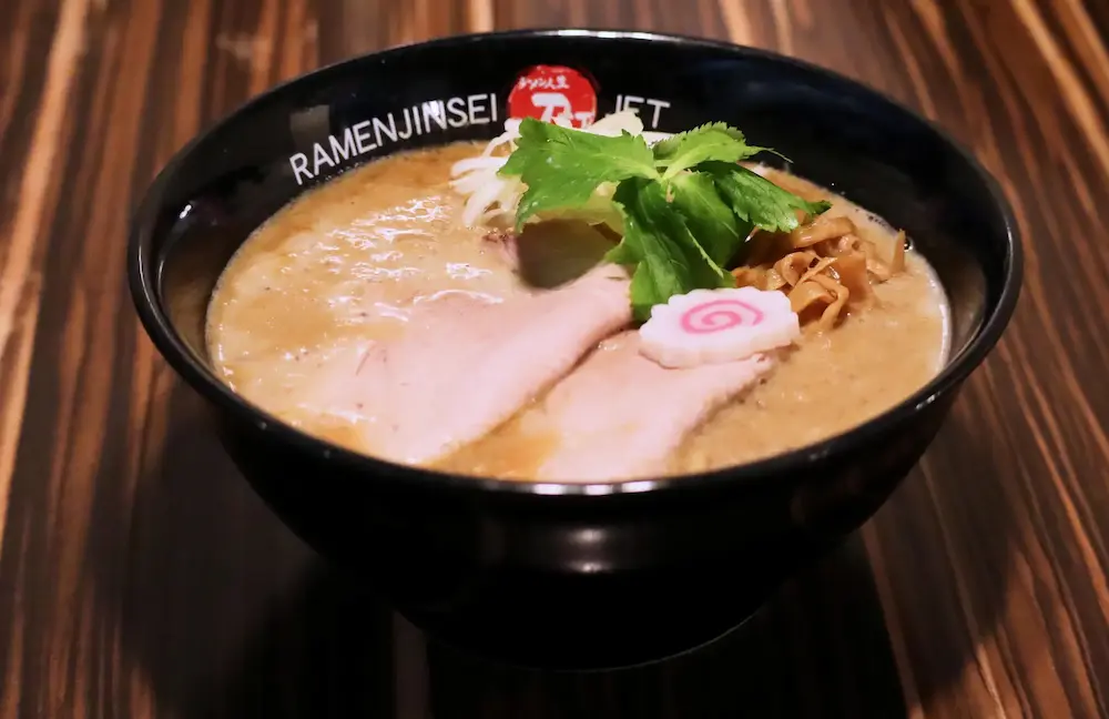 ラーメン人生JETで提供される濃厚鶏白湯ラーメンの実物写真。チャーシュー、なると、香草、メンマがのった一杯で、濃厚でコク深いスープが特徴。